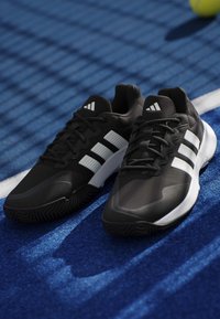 Αθλητικά παπούτσια Adidas σε ασπρόμαυρο, τοποθετημένα σε μπλε γήπεδο τένις κοντά σε λευκή γραμμή με μια θολωμένη μπάλα του τένις στο φόντο.