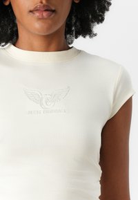 Kremfarget tettsittende t-skjorte med korte ermer, med forhøyet "GUESS ORIGINALS" logo og vingemønster på forsiden. Slett tekstur.