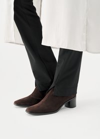 Καφέ σουέντ μποτάκια αστραγάλου με μυτερές μύτες και χοντρά τακούνια, σε συνδυασμό με μαύρο εφαρμοστό παντελόνι που ανοίγει ελαφρώς στο τελείωμα.