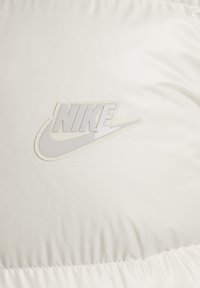Chaqueta acolchada blanca de Nike con un parche de logo reflectante, textura suave y dobladillo fruncido. La tela es ligera y resistente al agua.