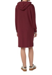 Bordeaux-rood hoodie jurk met lange mouwen, zijsplitten en contrasterende gestreepte accenten op de manchetten. Model gecombineerd met casual sneakers.