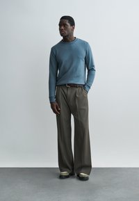 Mees seisab ühevärvilise seina ees, kandes sinist pikkade varrukatega särki, pruune laia säärega pükse vööga ja vabaajajalatsid.