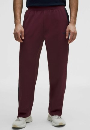 Man draagt wijnrode rechte trainingsbroek met zwarte zijstrepen en witte sneakers, staat tegen een effen lichte achtergrond.
