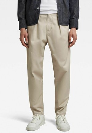 Homme portant un pantalon plissé beige, des baskets blanches, une chemise blanche et une veste en jean foncé sur fond blanc.