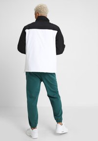 Svart og hvit jakke med høy krage, som har en ensfarget svart overdel og hvit underdel. Kombinert med grønne joggebukser og hvite joggesko.