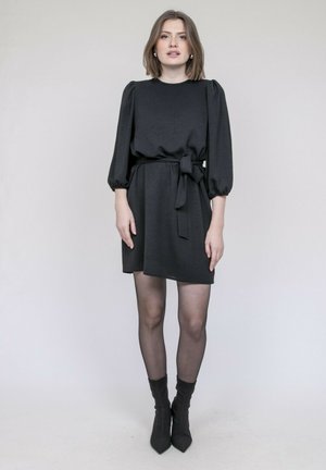 Robe noire avec manches bouffantes, texture en tissu, ceinture à la taille et longueur au-dessus du genou. Portée avec des collants transparents et des bottines pointues.