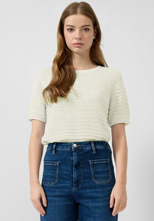 Młoda kobieta o długich brązowych włosach, ubrana w kremowy, teksturowany top z krótkim rękawem oraz wysoki stan niebieskich dżinsów, na jednolitym tle.