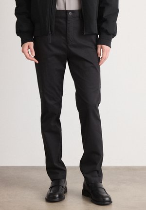 Mężczyzna w czarnych prostych spodniach i czarnych skórzanych mokasynach, stojący na tle jednolitego jasnego tła.