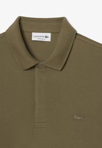 Olivgrön Lacoste polo med texturerad tygkvalitet, vikbar krage, tvåknappsknäppning och broderad logotyp på vänster bröst.