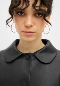 Schwarze Lederjacke mit rundem Kragen und Knopfverschlüssen. Das Model trägt große Ohringe. Glatte Textur und elegantes Design.