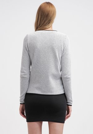 Femme aux longs cheveux blonds portant un haut blanc à manches longues avec un motif hexagonal noir et une jupe noire unie, debout de dos.