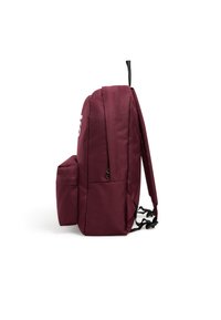 Un sac à dos en tissu marron présente une poche latérale, des bretelles noires réglables et une poignée de saisie en haut, avec un design simple et un détail logo.