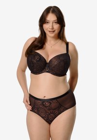 Kobieta w rozmiarze plus z długimi brązowymi włosami, ubrana w czarny koronkowy stanik i dopasowane majtki, uśmiechająca się na tle jednolitego białego tła.