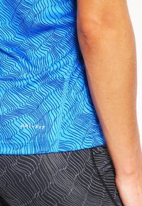 Gros plan sur une personne portant un t-shirt Dri-FIT bleu à motifs et un short de sport noir à motifs, montrant son bras droit et le côté du torse.