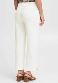 Pantalon blanc à jambe droite avec une taille élastique, fentes sur les côtés au niveau de l'ourlet, associé à des sandales à talons marron. Tissu lisse et léger.