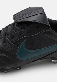 Crampons de football noires en cuir avec un design épuré, arborant un swoosh Nike teal et une languette texturée. Comprend un laçage traditionnel et des crampons.