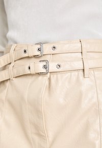 Ceinture en faux cuir beige avec deux sangles réglables, ferroneries en argent et trous renforcés le long de la longueur pour un ajustement sécurisé.
