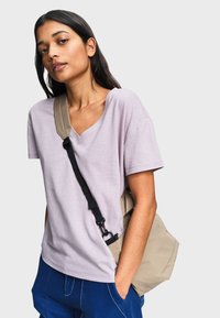 Jeune femme portant un t-shirt à col en V violet clair, un pantalon bleu et un sac bandoulière beige, posant devant un fond uni.