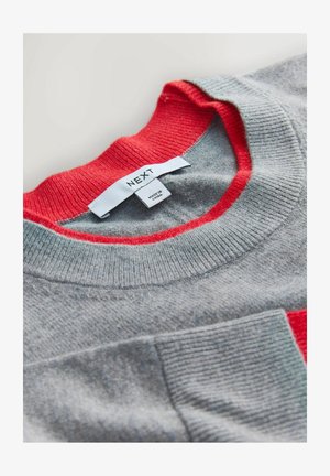 Šedý svetr s červeným žebrovaným límcem a kapsou. Štítek s názvem "NEXT" uvádí informace o péči o tkaninu a původu. Měkká textura.