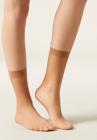 Nude ankelstrømper med en blød, tekstureret overflade og elastiske topkanter, der hviler på bare fødder. Det ene fod vist fra siden, den anden frontalt.