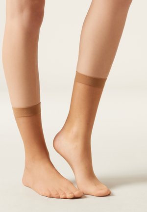 Nude ankelsockor med en mjuk, texturerad yta och elastiska toppband, vilande på nakna fötter. En fot visas från sidan, en från framsidan.