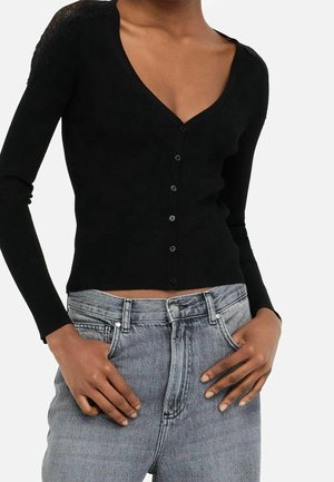 Personne portant un haut noir à manches longues boutonné en V avec des détails en dentelle sur les épaules et un jean gris clair taille haute, les mains dans les poches.