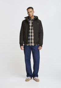 Schwarze Kapuzenjacke über einem karierten Hemd, lockere blaue Jeans und beigefarbene Sneakers. Glatte Textur mit einer seitlichen Reißverschlusstasche an der Jacke.