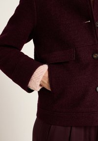 Hand in pocket of dark burgundy wool jacket, pink knitted sweater cuff visible at wrist, paired with matching burgundy trousers.