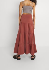 Jupe maxi en tissu doux couleur rouille, présentant un design à volants avec des accents froncés. Associée à un débardeur gris court et des sandales noires.