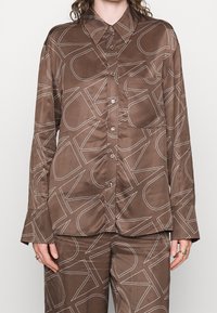 Ruskea satiininen nappipusero ja siihen sopivat housut, joissa on suuri valkoinen reunakirjaimisto, päällä henkilö, jolla on sormuksia sormissa.