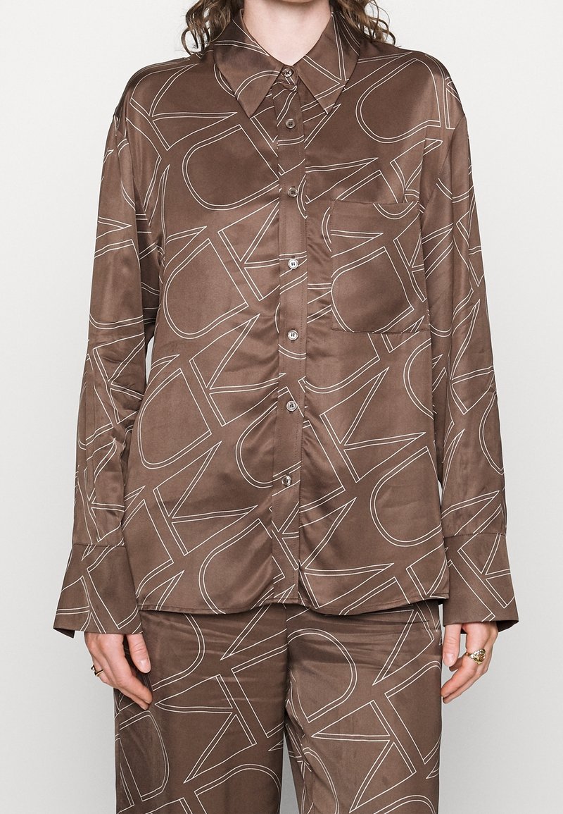 Ruskea satiininen nappipusero ja siihen sopivat housut, joissa on suuri valkoinen reunakirjaimisto, päällä henkilö, jolla on sormuksia sormissa.