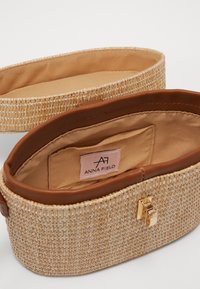 Borsa ovale realizzata in materiale intrecciato simile alla paglia con dettagli in pelle marrone. L'interno presenta una fodera beige e un etichetta con logo. Dettaglio con chiusura dorata.