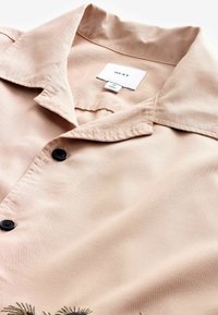 Koszula w kolorze beżowym z krótkim rękawem, wykonana z miękkiego materiału, z kołnierzykiem rozłożystym, ozdobiona czarnymi guzikami i haftowanymi detalami w kwiaty.