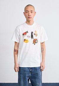White cotton t-shirt featuring colorful prints of dogs, a hand, a disc, and a pool float. The fit is relaxed with short sleeves.