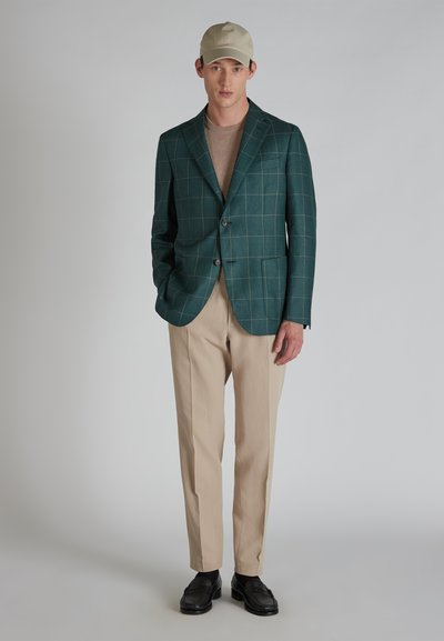 Groen geruite blazer met twee knopen, beige trui eronder, beige broek en zwarte schoenen; een eenvoudige pet maakt de outfit compleet.