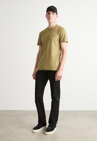 Olivově zelené tričko s krátkým rukávem a kulatým výstřihem, spárované s černými slim fit džíny a černými teniskami s bílou podešví. Nosit černou kšiltovku.