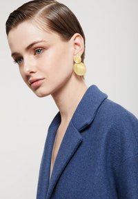 Veľké, textúrované zlaté náušnice s okrúhlymi tvarmi; nosené s modrým kabátom s golierom, zobrazujúce detailný záber na doplnok a dekolt.