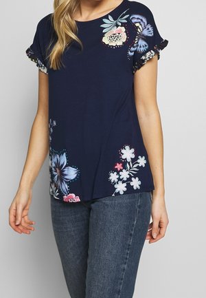 Granatowy T-shirt z krótkim rękawem z miękkiej tkaniny, zdobiony kwiatowymi nadrukami na przodzie i falbankami na mankietach jako detal. Standardowy krój z okrągłym dekoltem.