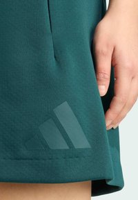 Green athletic skirt made of soft fabric, featuring a stitched hem and a translucent Adidas logo on the lower left side.