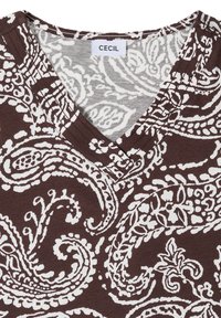 Chemise marron à motifs cachemire et floraux blancs et col en V avec l’étiquette "Cecil" à l’encolure.