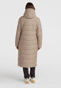 Lång beige pufferjacka med huva, med horisontellt quiltade sektioner, som når mitt på vaden, i kombination med svarta byxor och vita sneakers.