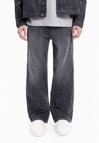 Szare dżinsowe spodnie o szerokich nogawkach wyróżniają się kontrastowym przeszyciem w zygzak i srebrnymi detalami metalowymi. Materiał ma teksturowane wykończenie z widocznymi szwami.
