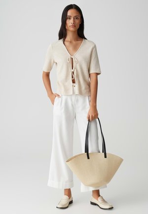 Knit beige top with a V-neck and tie closure, paired with white wide-leg pants. The model carries a beige straw bag with black handles.