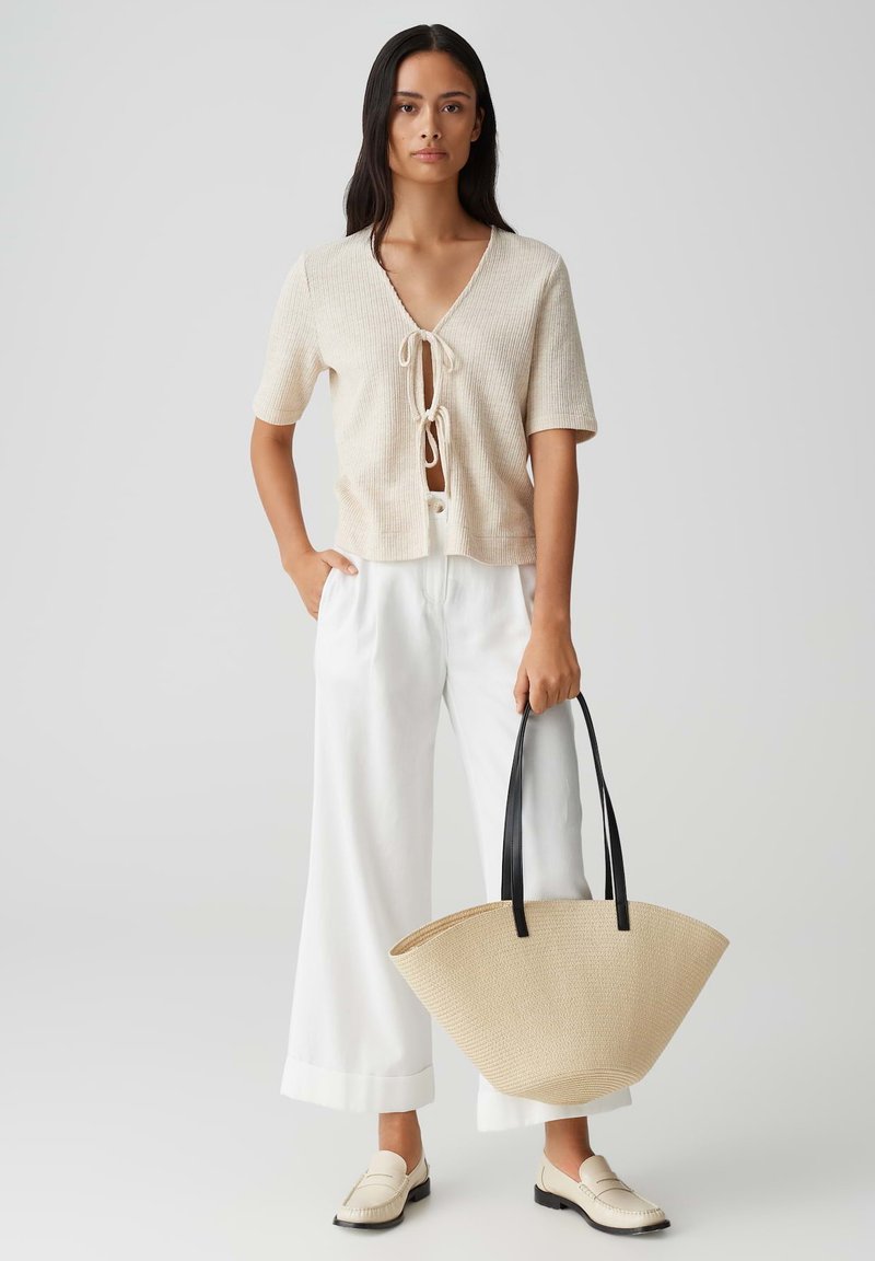 Top beige lavorato a maglia con scollatura a V e chiusura a fiocco, abbinato a pantaloni bianchi a gamba larga. La modella porta una borsa di paglia beige con manici neri.