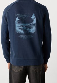 Felpa della marina con maniche lunghe, caratterizzata da una grafica di un paesaggio blu e testo sul retro. Tessuto morbido con orlo e polsini a coste.