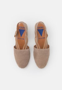 Espadrilles de ante beige con tiras elásticas, puntera redonda y suela de yute. Presentan un logo en forma de triángulo azul en el interior, etiquetado "VERBENAS."