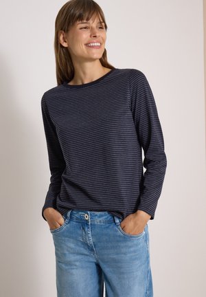 Femme souriante portant un t-shirt à manches longues rayé bleu marine et un jean bleu clair, les mains dans les poches, sur un fond uni.