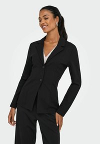 Femme aux cheveux foncés attachés en queue de cheval, portant un blazer et un pantalon noirs sur mesure par-dessus un haut blanc, souriant les yeux fermés devant un fond uni.