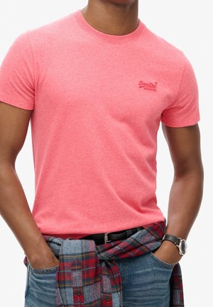 Mężczyzna ubrany w dopasowany, koralowo-różowy T-shirt z krótkim rękawem i małym logo, niebieskie dżinsy, koszula w kratę związana wokół talii oraz czarny zegarek na nadgarstku.