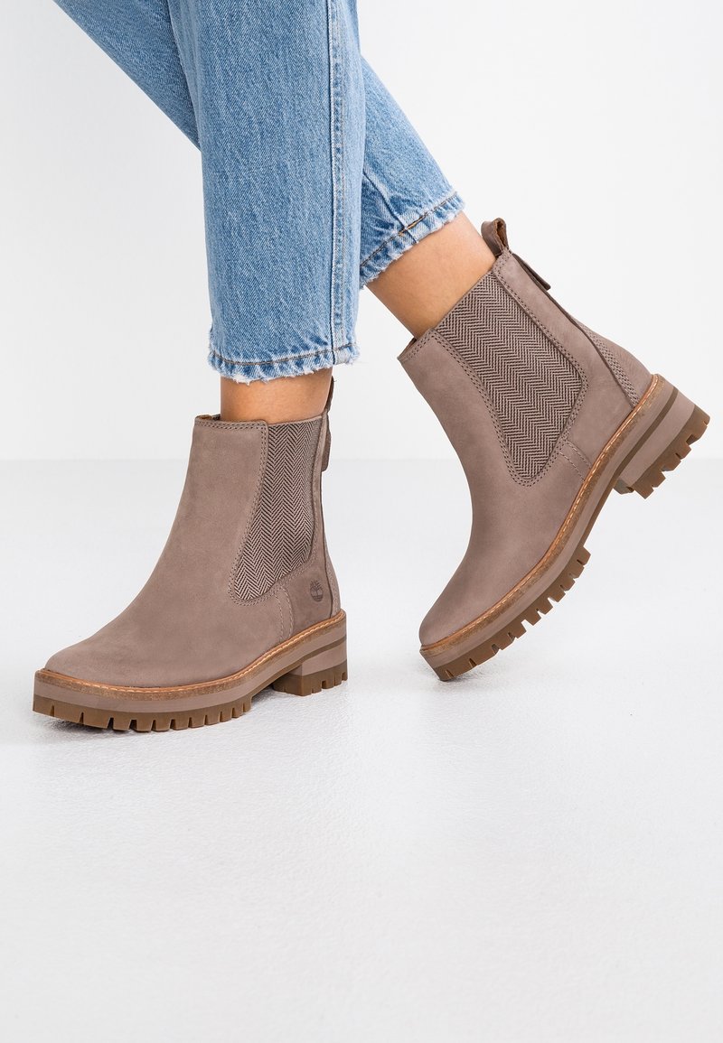 Bottes marron en daim montant jusqu'à la cheville avec des panneaux latéraux élastiques texturés, portées avec un jean bleu clair raccourci sur un sol blanc.